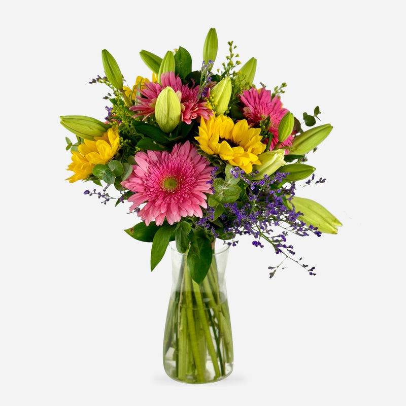 Ramo Alegría con gerberas rosadas, girasoles amarillos, capullos de lirio y flores moradas en jarrón de cristal.