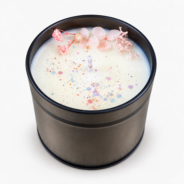 Vela aromática de soja en recipiente oscuro, con flores decorativas rosas y mecha sin encender sobre fondo neutro.
